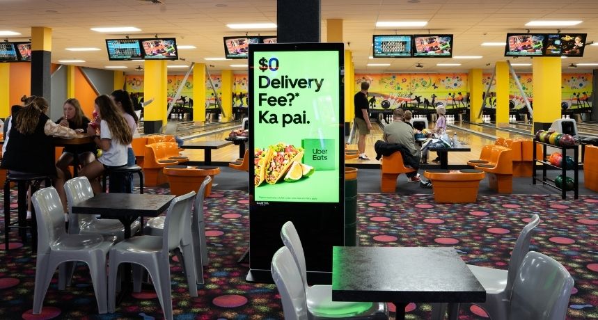 A digital advertising screen at Bowlarama New Plymouth with bowling lanes and people in background